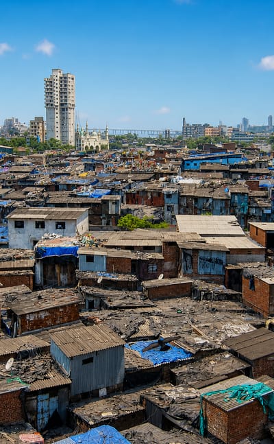 The Dharavi settlement in Mumbai with dense corrugated-roof shanties and blue tarps below distant high-rise towers