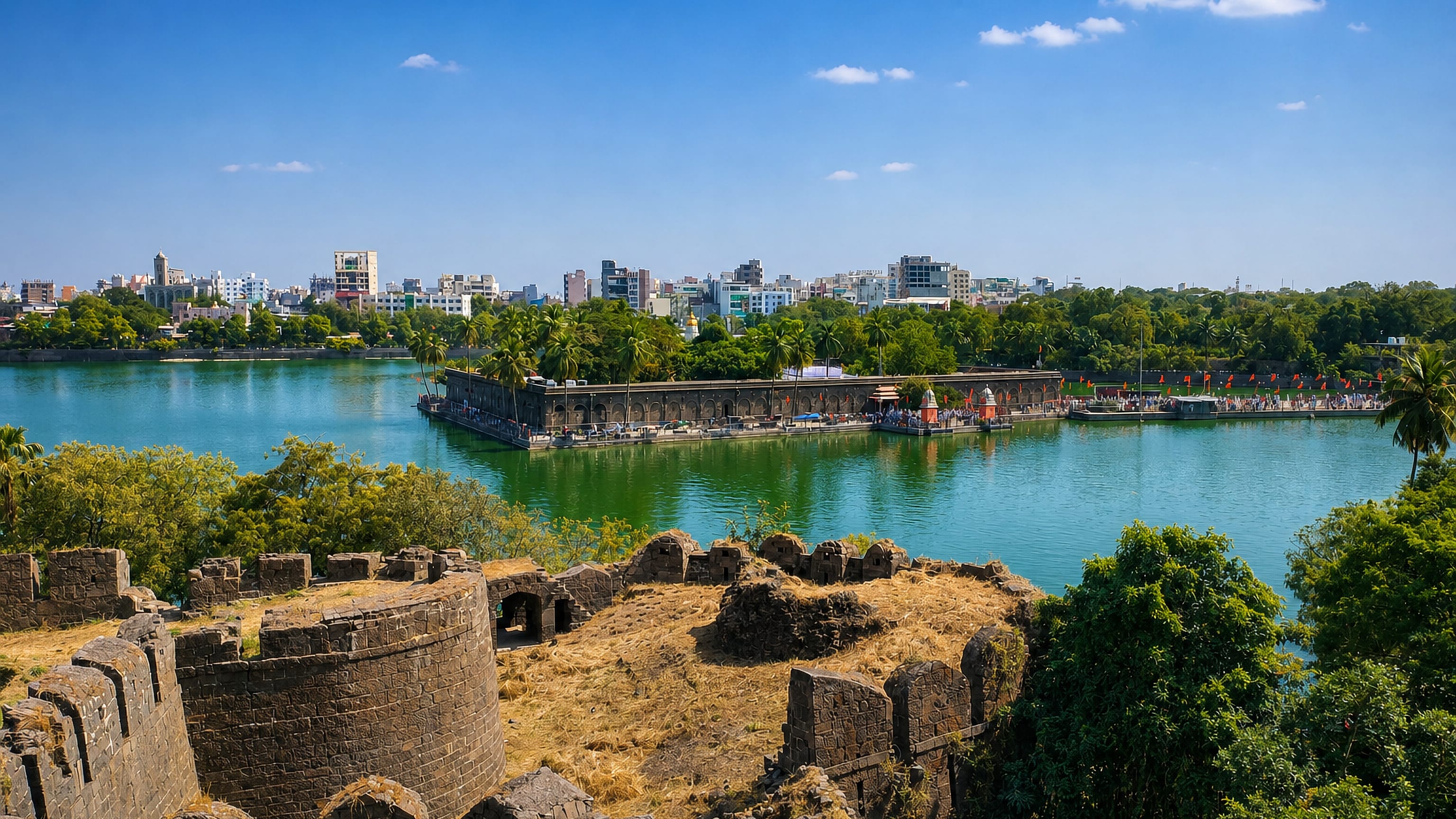 Stone fort ramparts above a green lake in Sholapur, Maharashtra, with an island temple complex and city skyline.