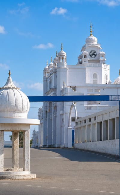 White domed gurdwara complex with a clock tower and gateway arch beside an open road in Sirhind, Punjab.