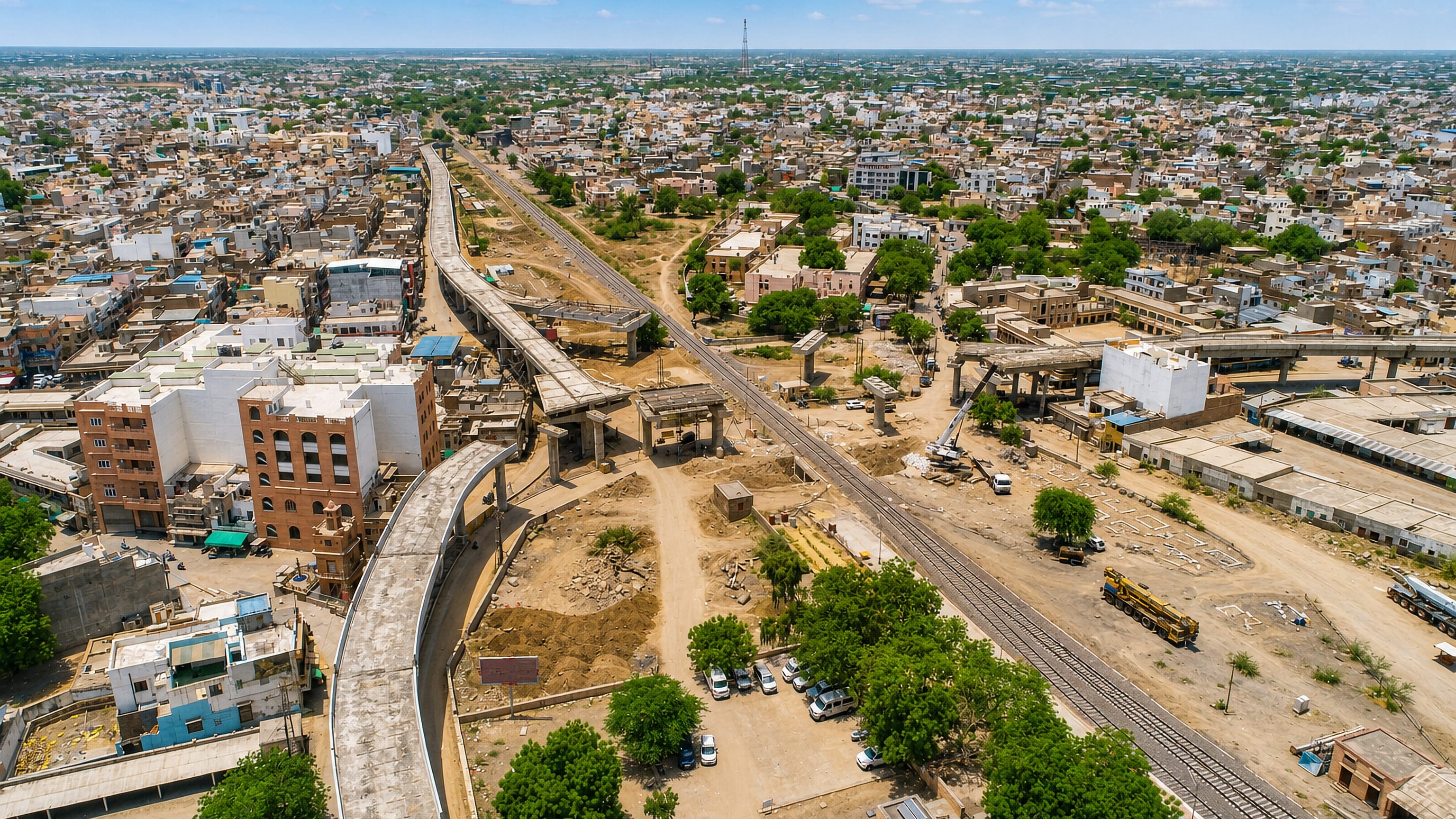 Aerial view over Balotra, Rajasthan, showing dense rooftops, railway tracks and an elevated flyover under construction.