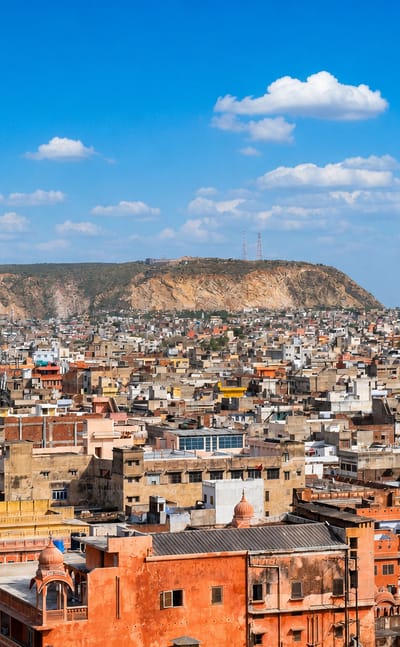 Dense pink-hued rooftops of Jaipur's old city spreading toward the surrounding Aravalli hills under a blue sky
