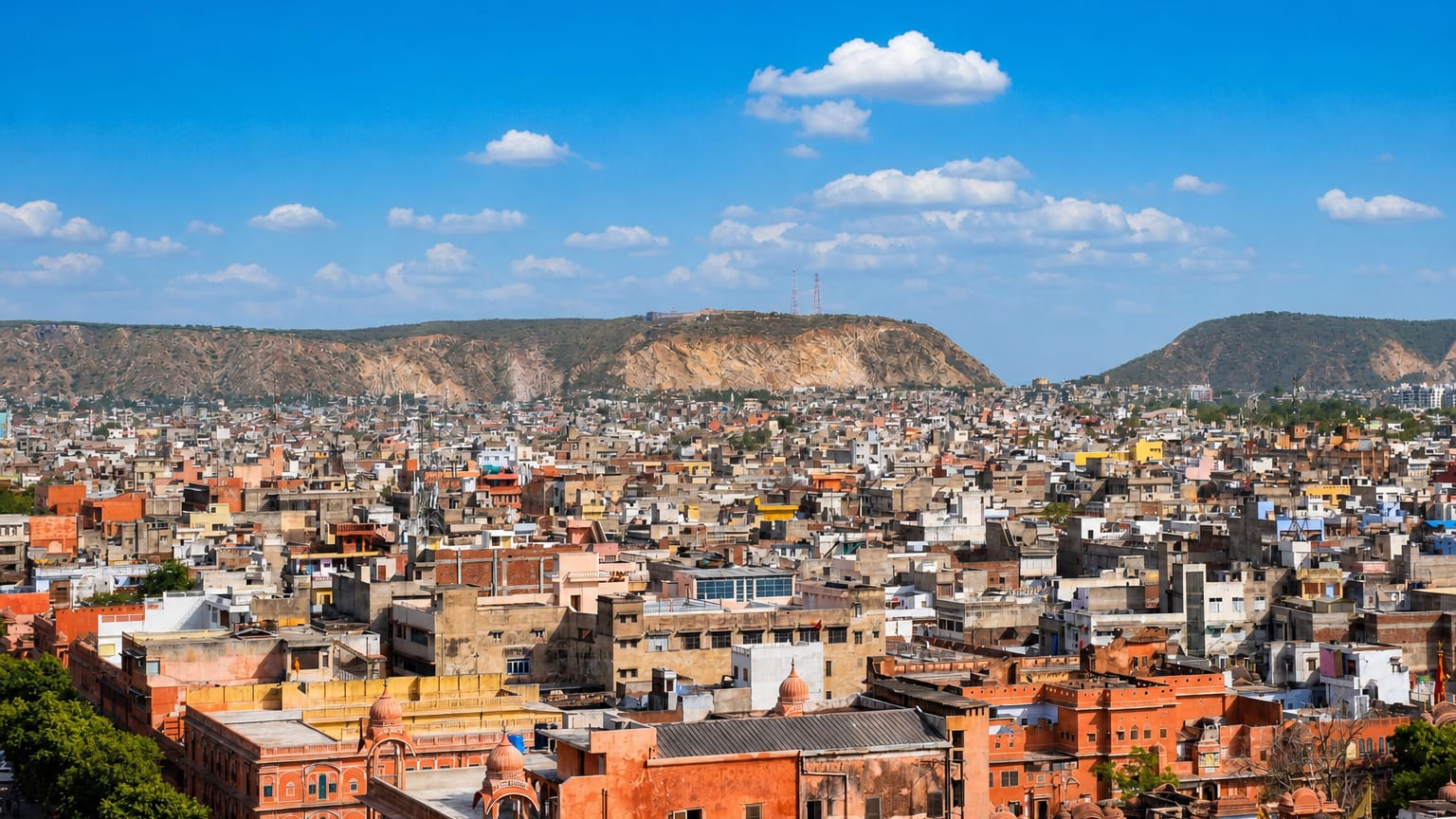 Dense pink-hued rooftops of Jaipur's old city spreading toward the surrounding Aravalli hills under a blue sky