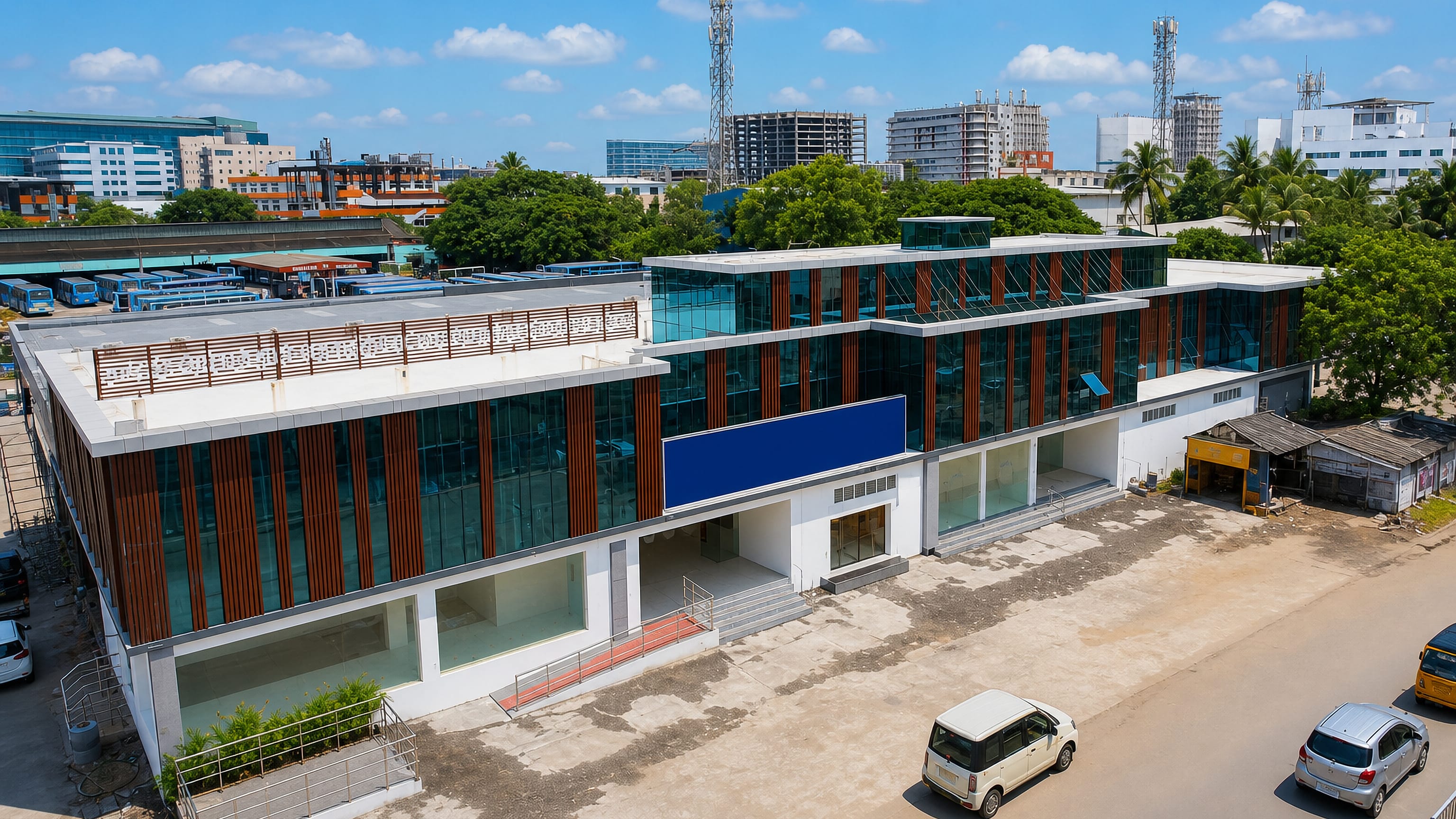 A modern glass-and-timber commercial block beside a bus depot in Ambattur, Chennai, under a bright sky.
