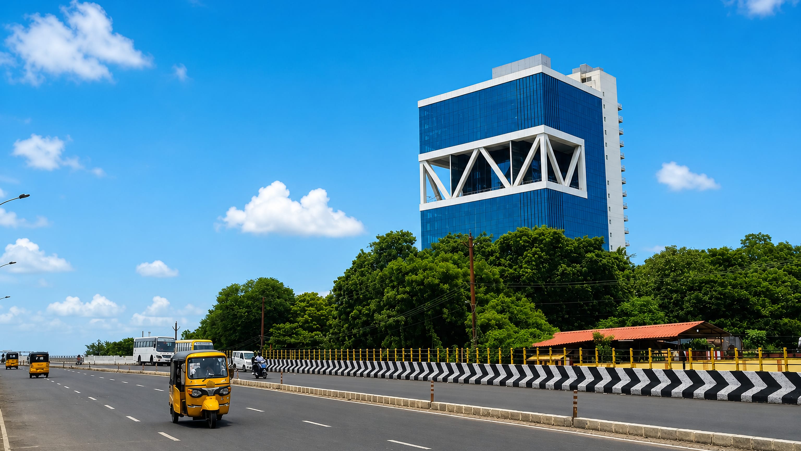 Wide highway with an auto-rickshaw passing a blue glass office tower in Avadi, Tamil Nadu, India.