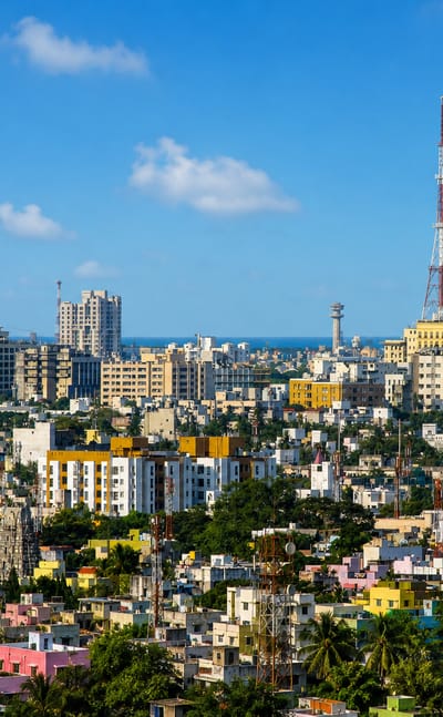 Chennai skyline with dense low-rise housing, palm trees and the Doordarshan lattice tower under a bright sky, sea on the horizon.
