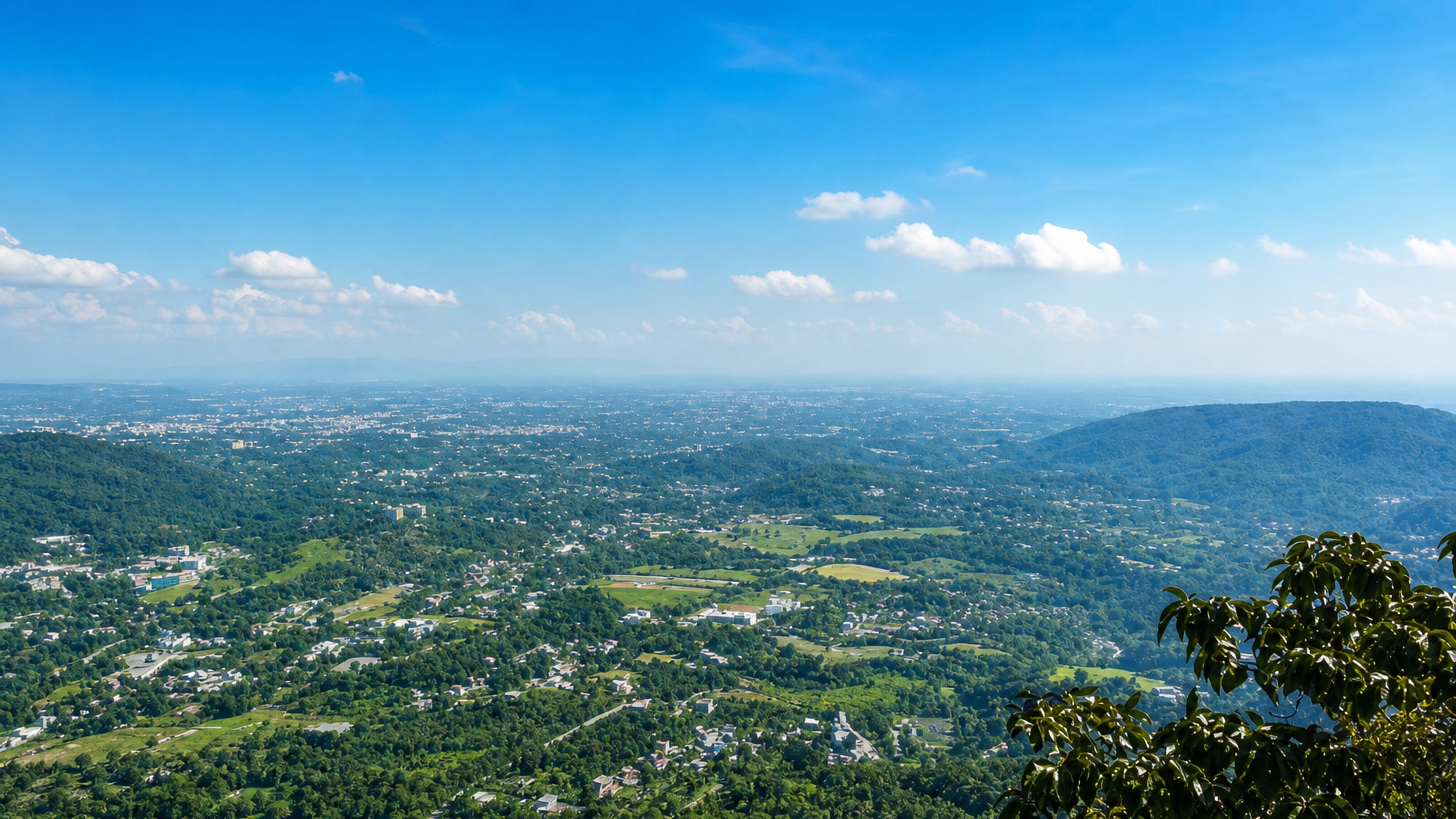 Dehradun spread across the Doon Valley below forested Himalayan foothills under a bright blue sky.