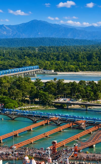 Bridges cross the turquoise Ganges past Haridwar's riverside ghats with Himalayan foothills beyond.