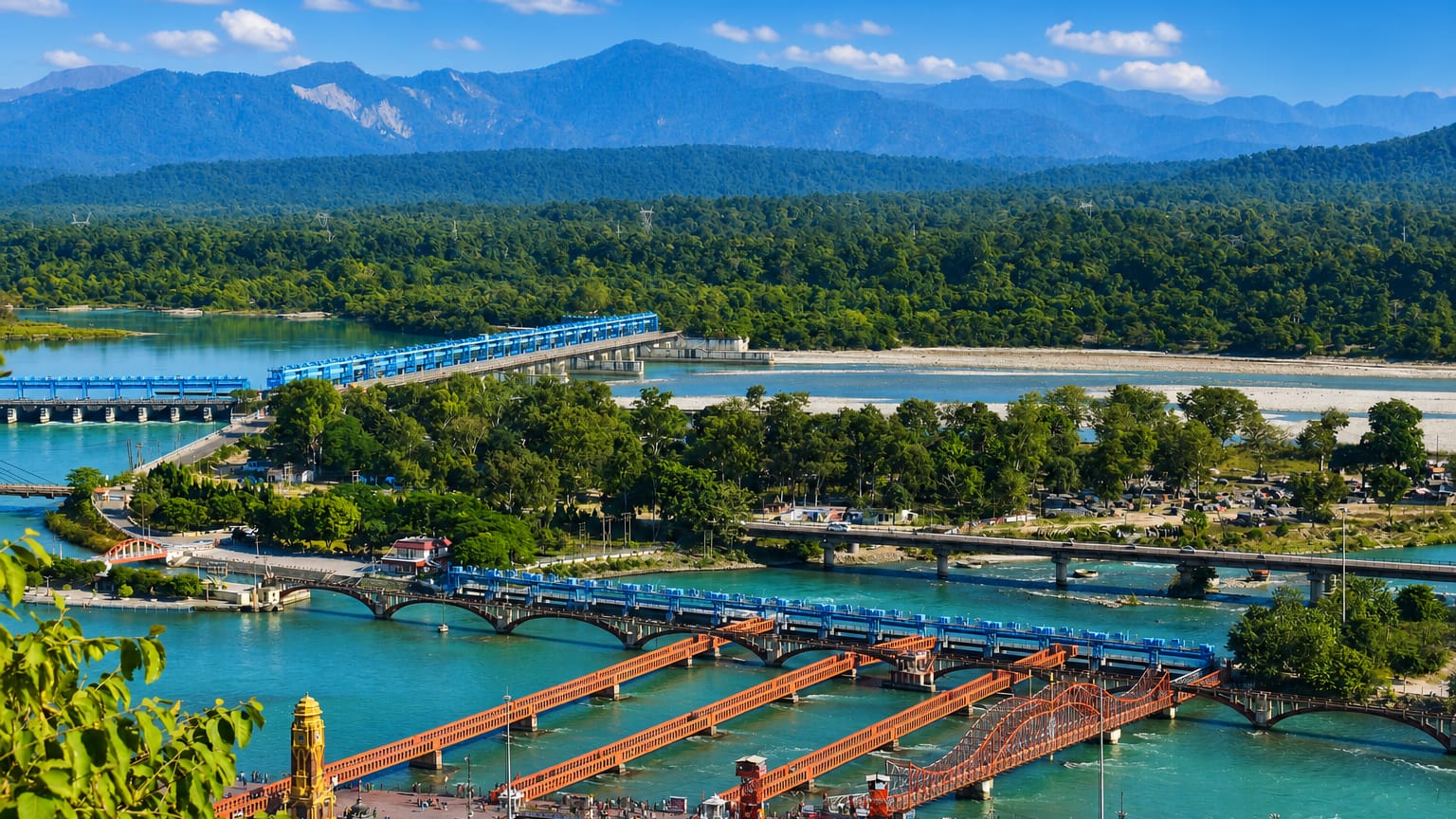 Bridges cross the turquoise Ganges past Haridwar's riverside ghats with Himalayan foothills beyond.