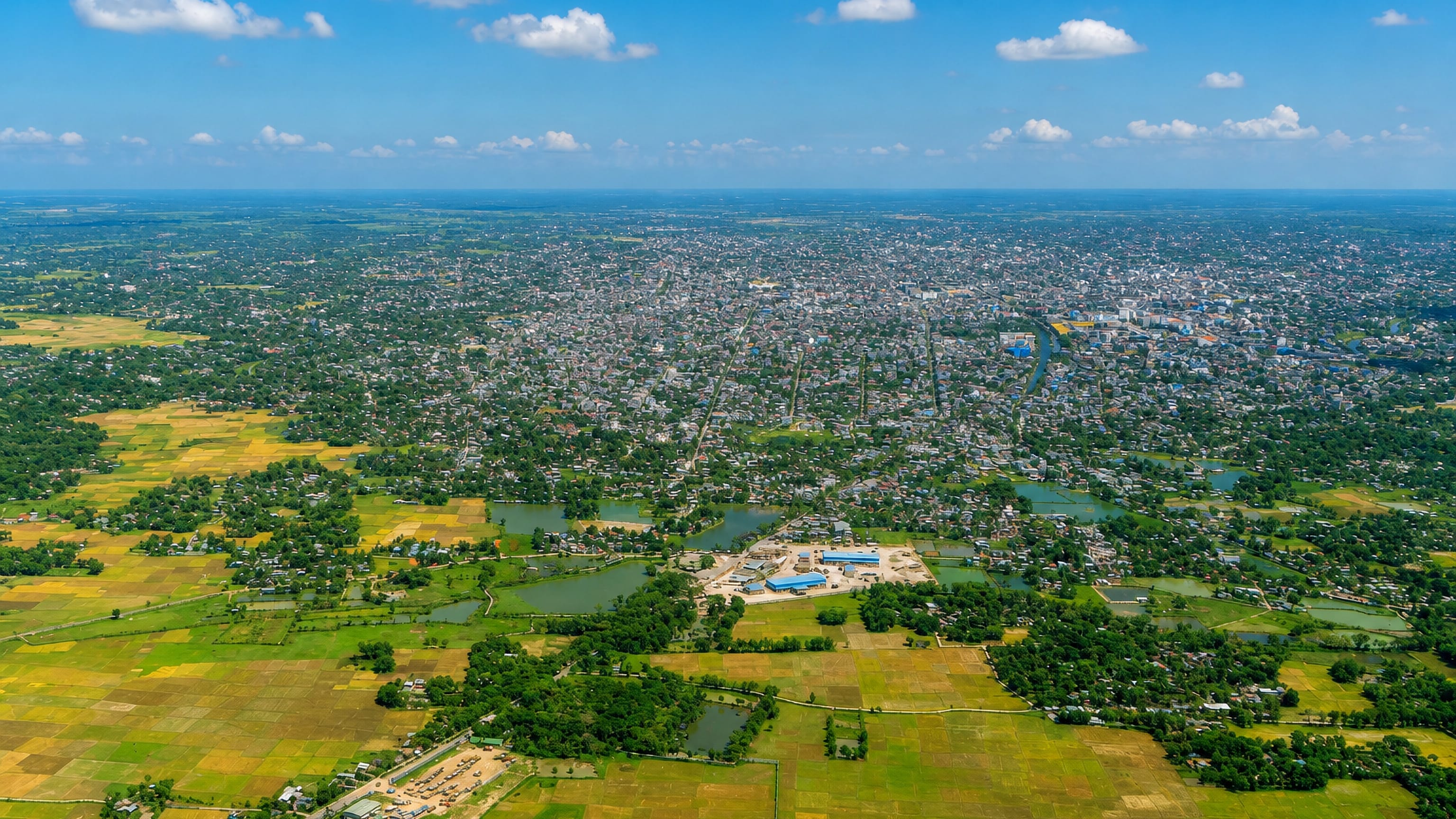 Agartala's dense low-rise sprawl seen from the air, ringed by paddy fields and ponds under a bright sky