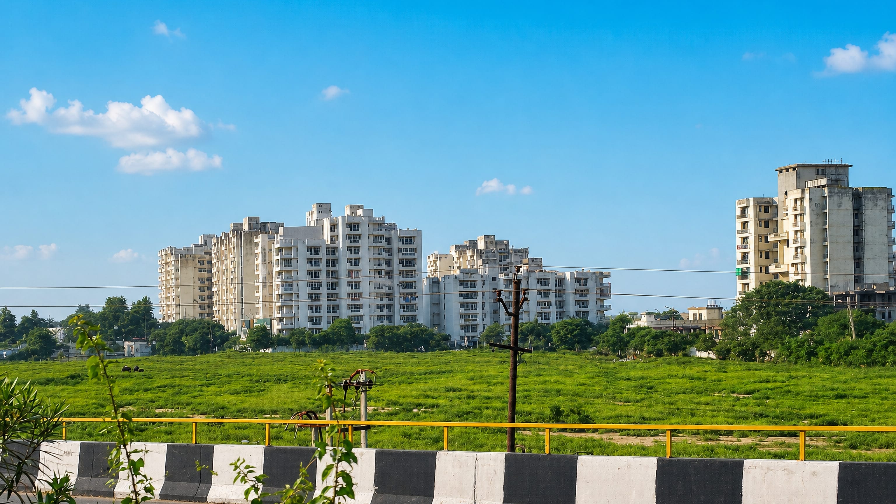 Whitewashed apartment towers rising beyond a green field along a roadside in Morādābād, India.