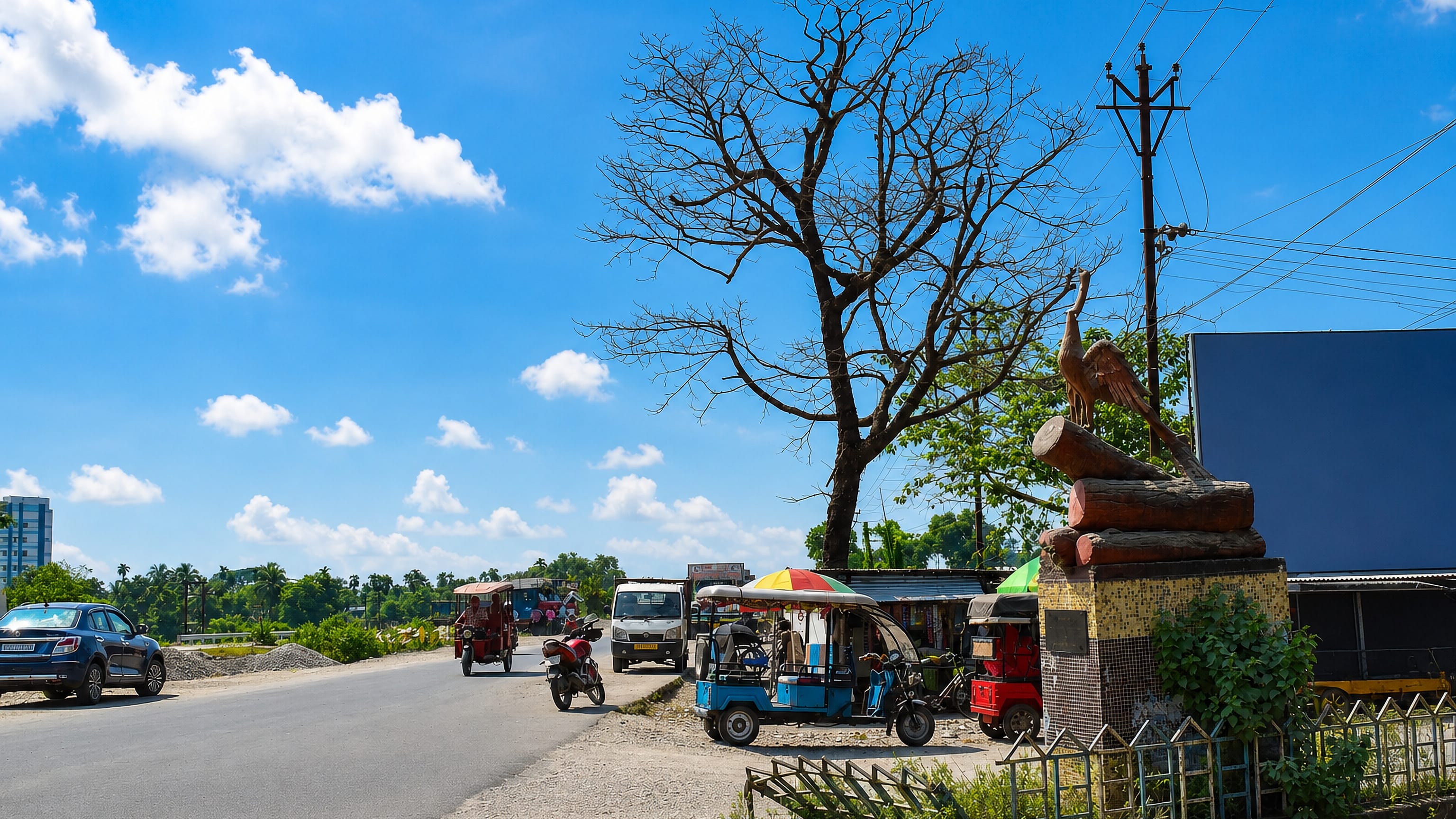 Roadside scene in Jalpaiguri, India, with e-rickshaws, stalls and a bird statue under a bare tree.
