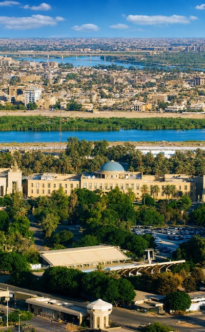 Aerial view of Baghdad with the Tigris River curving past green banks and a sprawling low-rise city under clear skies