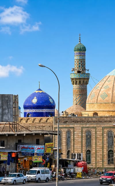 Ornate mosque with tiled domes and twin minarets above a busy market street in Basra, Iraq.