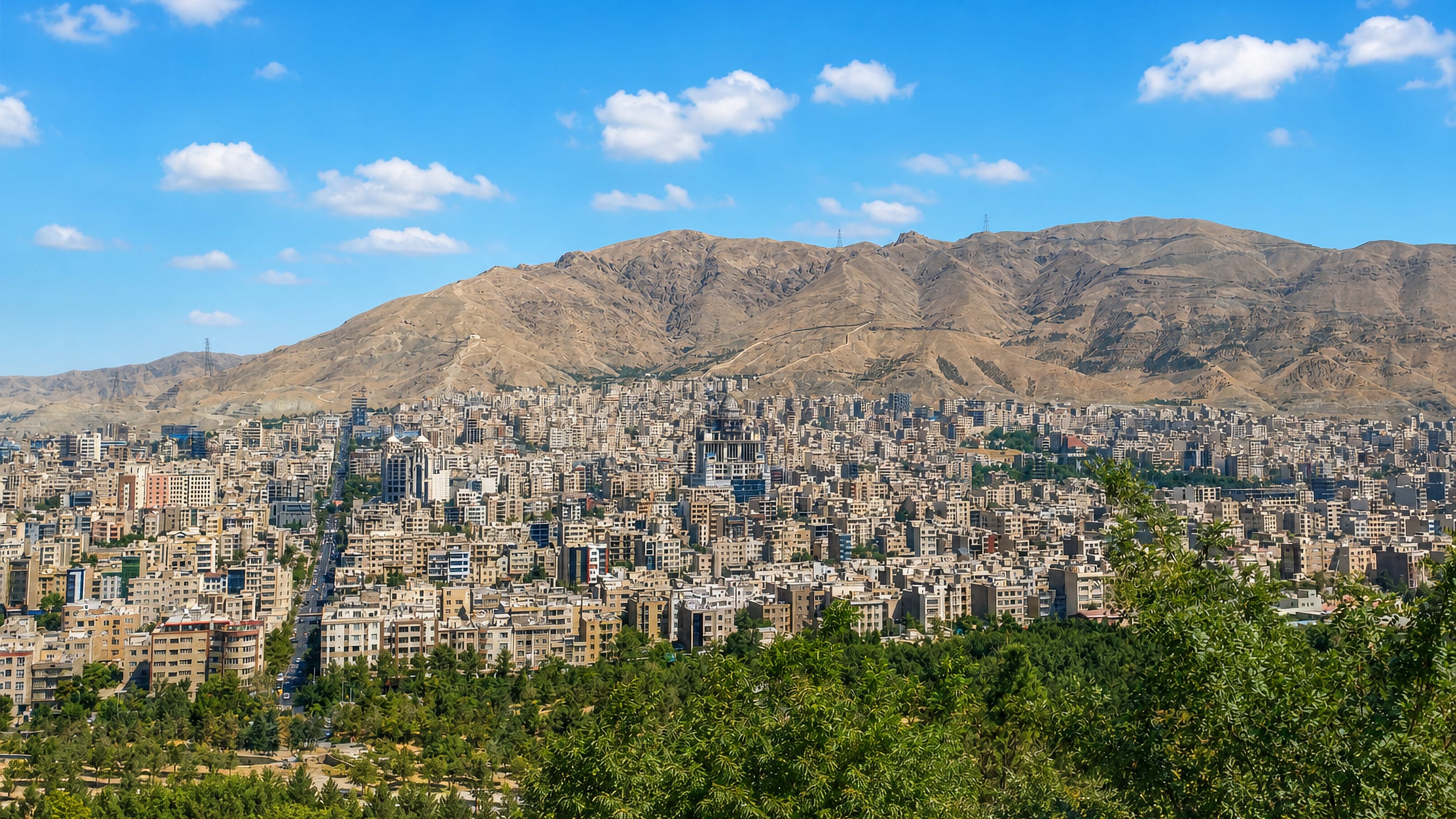 Dense beige apartment blocks of Karaj spreading below the arid Alborz foothills under a clear blue sky.