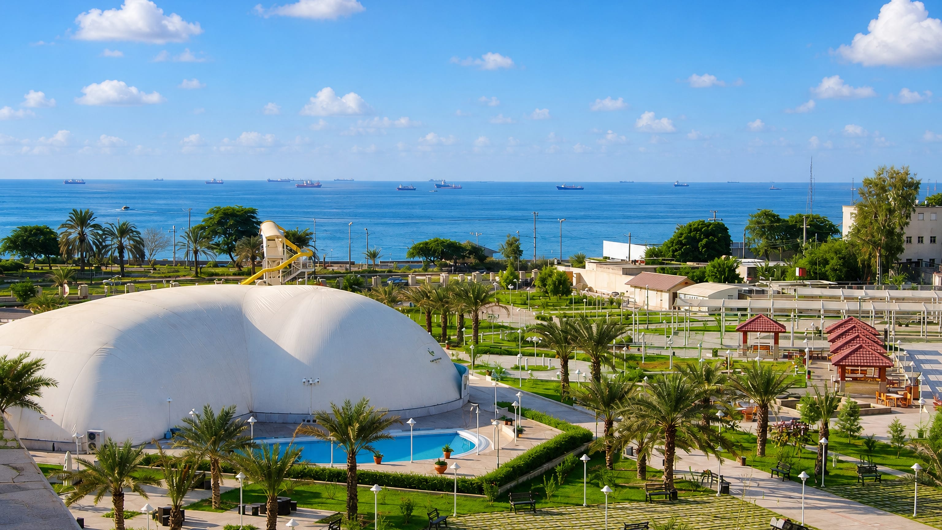 A palm-lined seafront park in Bandar Abbas with cargo ships anchored offshore in the Persian Gulf.