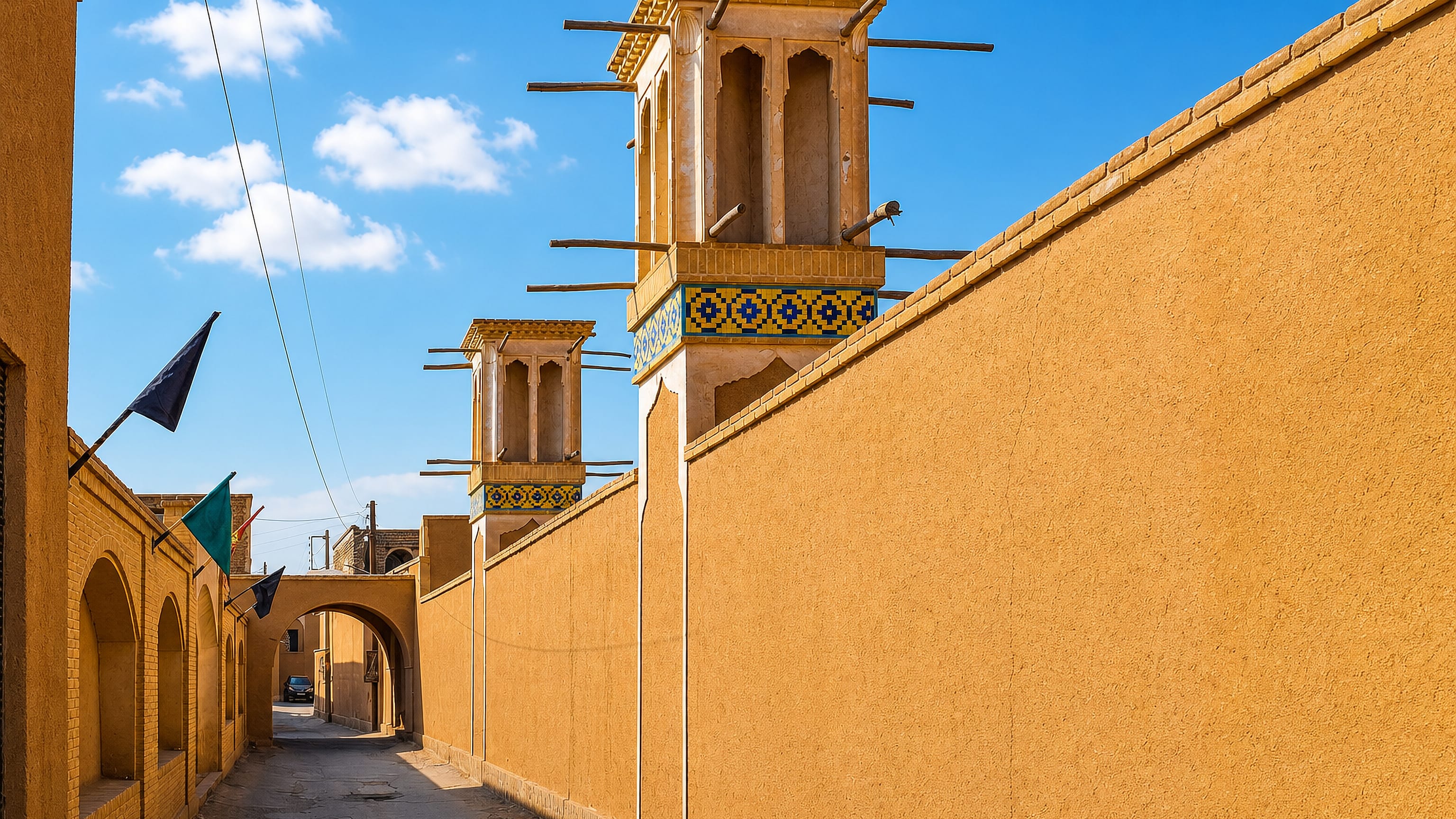 Mud-brick lane in Ārān Bīdgol lined by high desert walls and tiled wind-catcher towers under blue sky