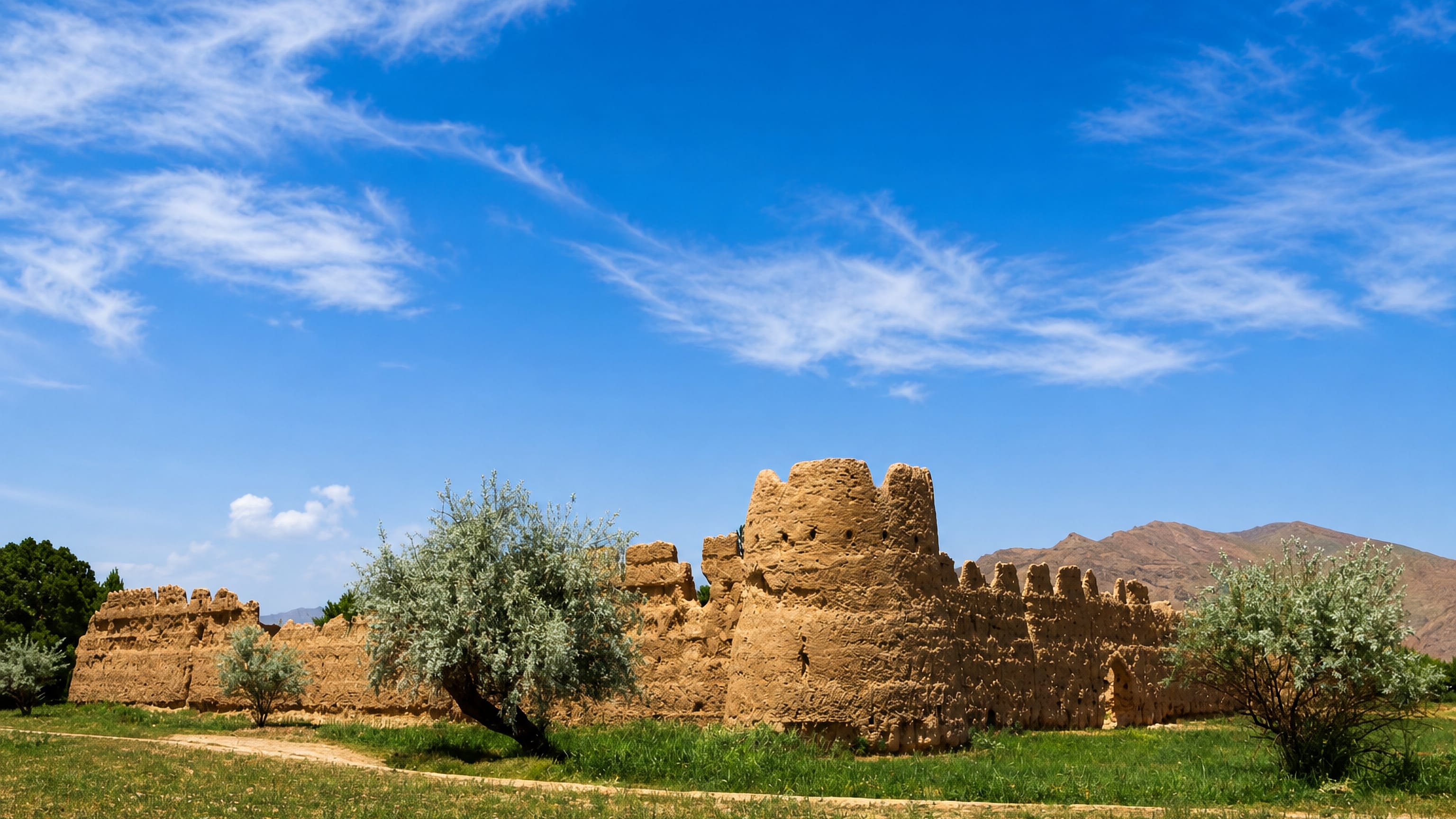 Mud-brick fortress walls standing in green fields below dry mountains near Golpāyegān, Iran.