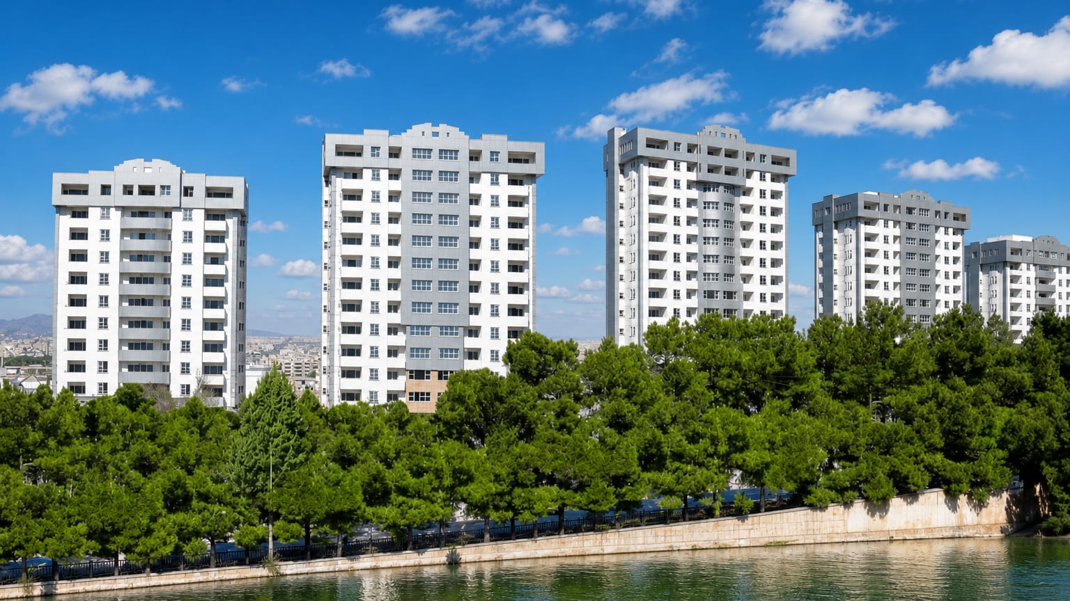 Modern white residential towers above pine trees along a riverbank in Isfahan, Iran.