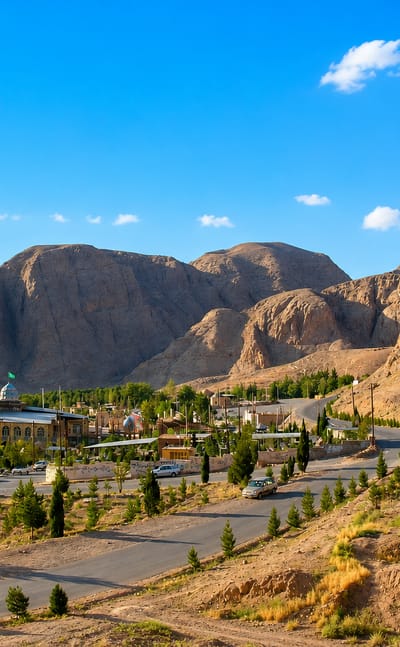 Kerman's desert edge with a blue-domed mosque and minaret below rugged bare mountains and cypress trees