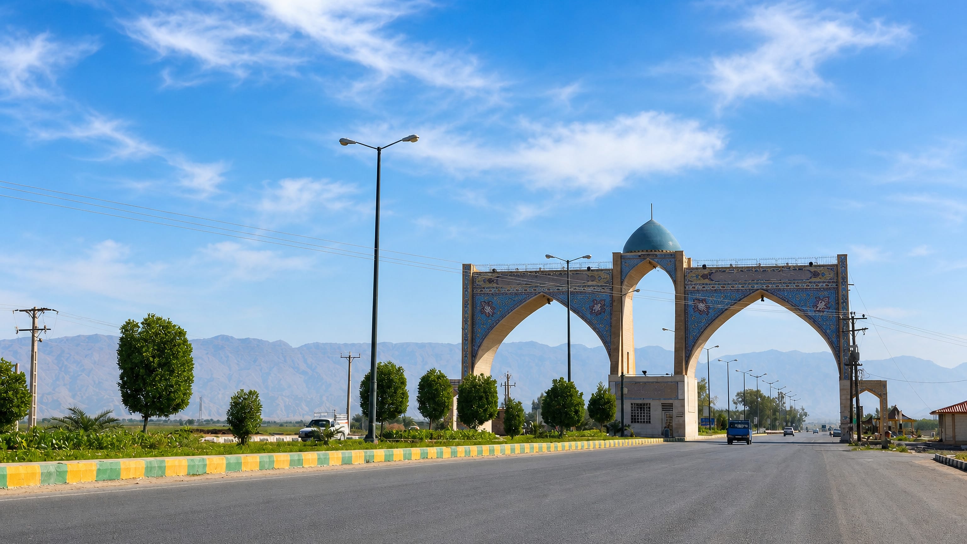 Tiled entrance gateway with a turquoise dome on the highway into Behbahān, Iran, with mountain foothills behind.