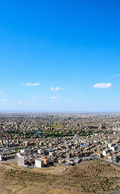 Sprawling low-rise cityscape of Qom seen from surrounding desert hills under a blue sky.