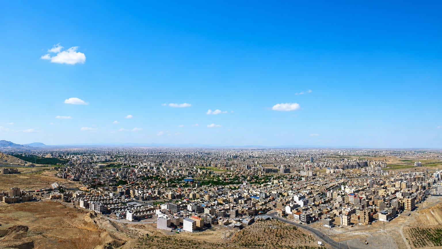 Sprawling low-rise cityscape of Qom seen from surrounding desert hills under a blue sky.