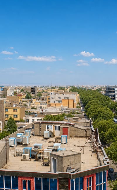 Rooftop view across Neyshabur's low-rise sunlit buildings toward a tree-lined boulevard under a clear blue sky