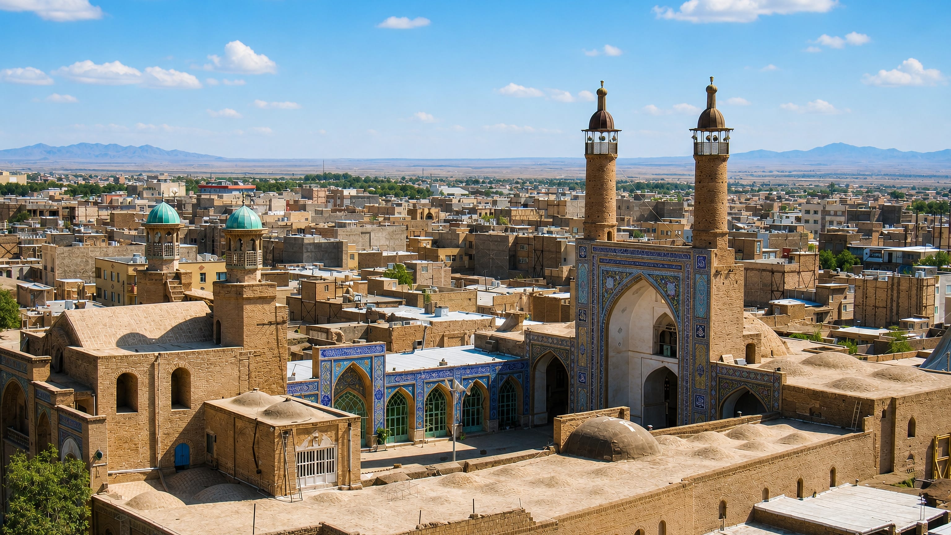 Mud-brick rooftops of Sabzevar, Iran, around a tiled congregational mosque with twin minarets below desert hills.