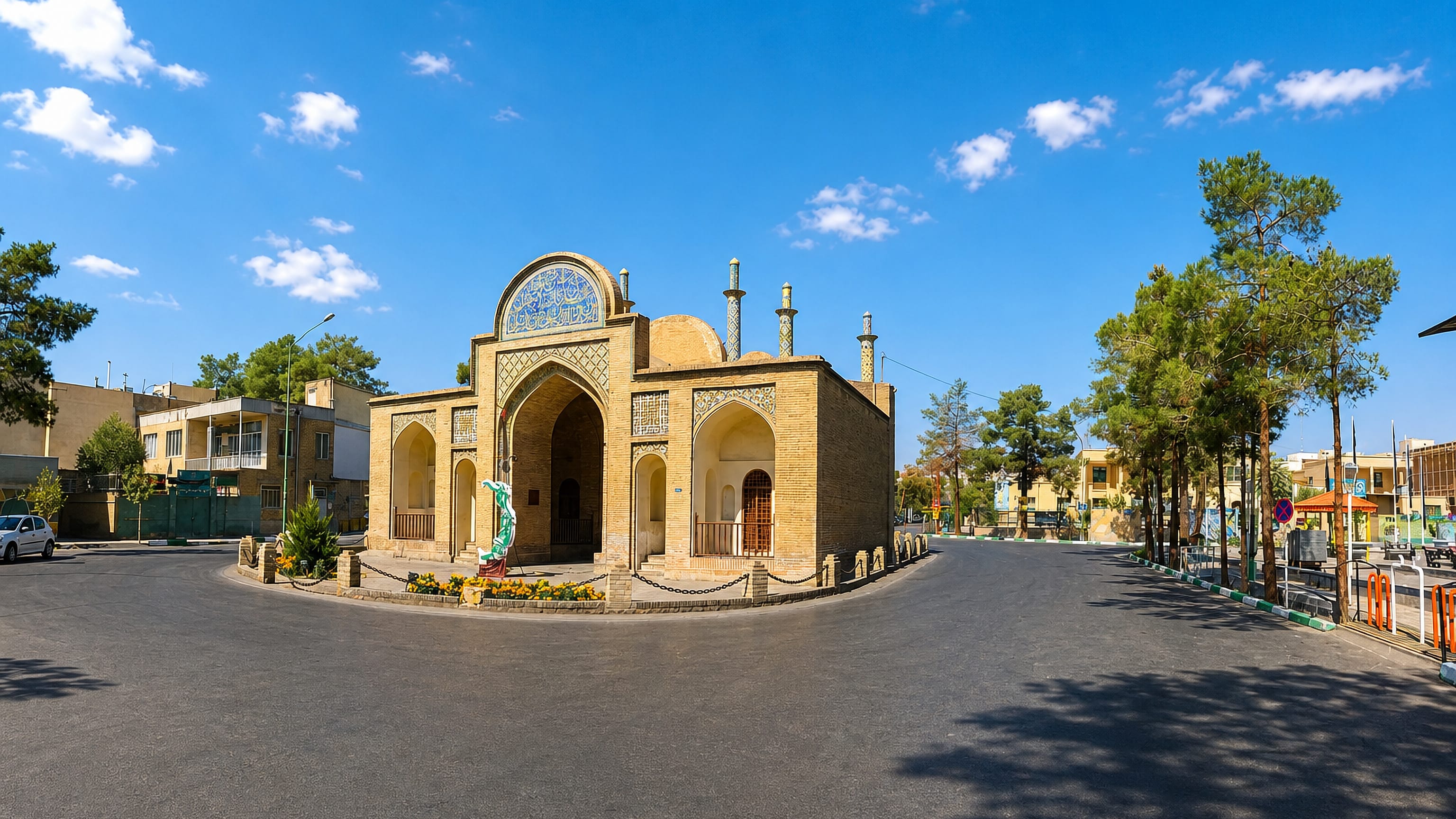 Tiled brick shrine with a domed iwan portal on a quiet roundabout in Semnan, Iran, under a clear sky.
