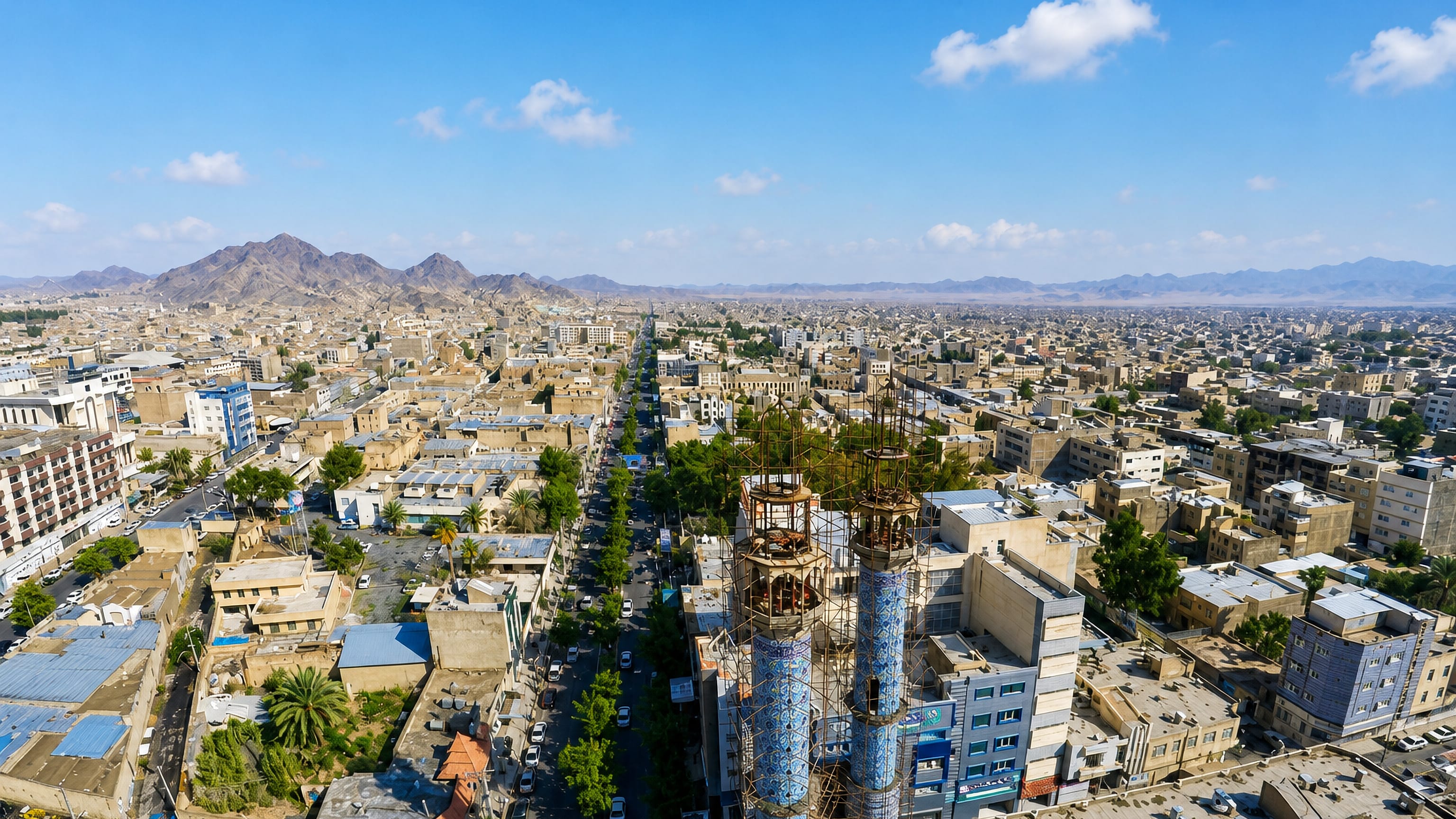 Aerial view of Zahedan, Iran, with tiled mosque minarets and dome above low desert blocks and arid hills.
