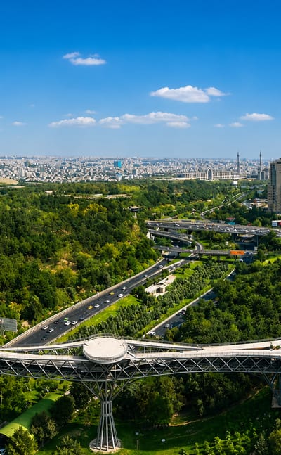 Tehran's Tabiat Bridge and green parkland in Abbas Abad, with the Iranian flag and city skyline beyond.