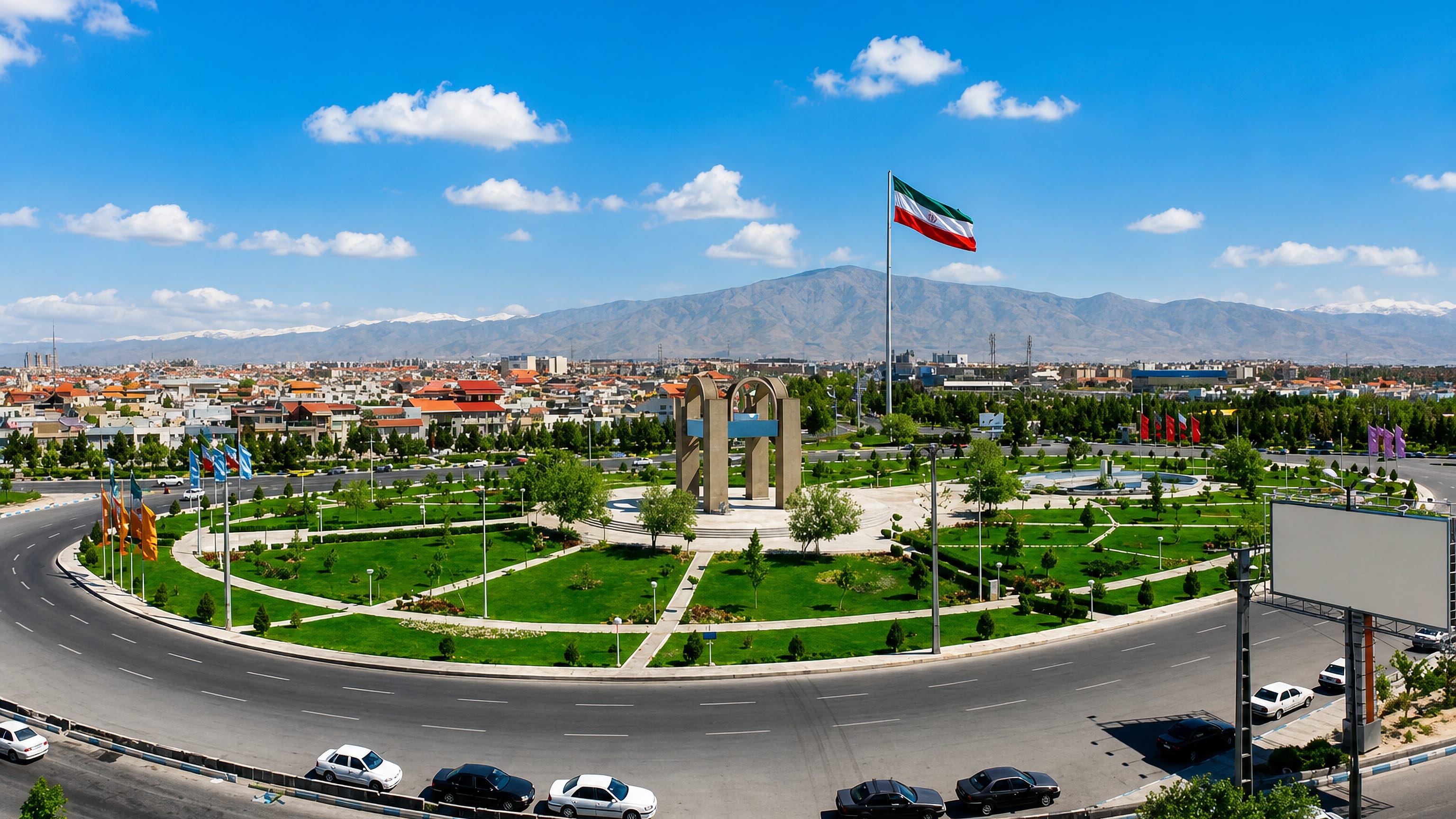 A landscaped roundabout with monument and Iranian flag in Andisheh, with the Alborz mountains behind.