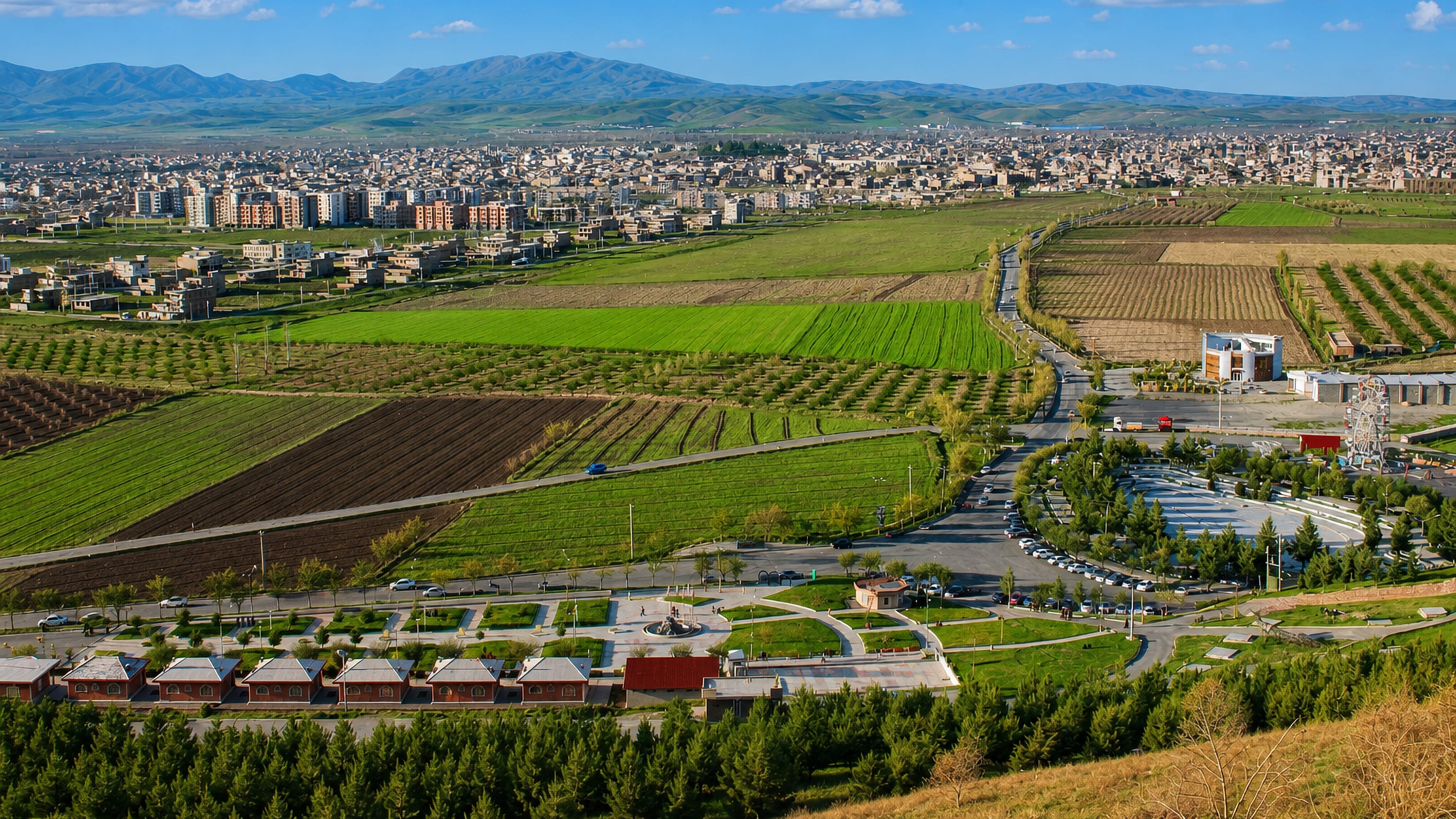 Hillside view over Naqadeh, Iran, with a landscaped park, green farm plots and mountain ridges beyond.