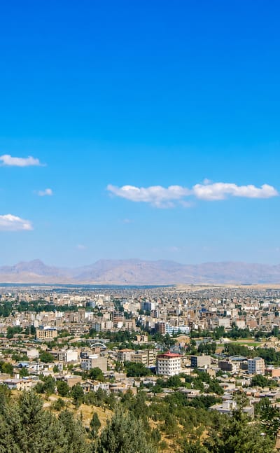 Orūmīyeh spread across a plain with distant mountains, seen from a green hillside under a clear blue sky.
