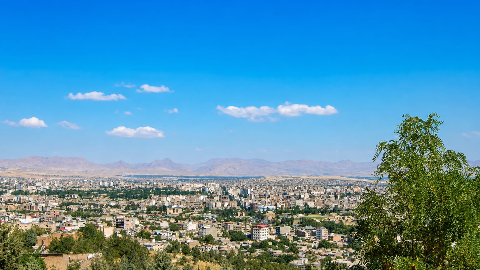 Orūmīyeh spread across a plain with distant mountains, seen from a green hillside under a clear blue sky.