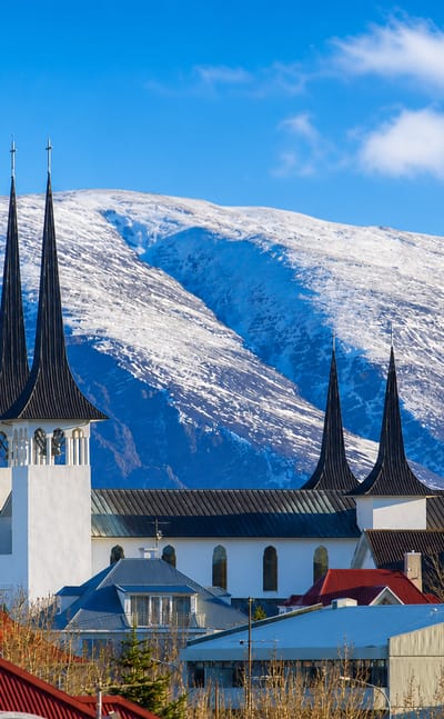 A white church with tall dark spires above Kópavogur, Iceland, set against snow-capped mountains and clear blue sky.