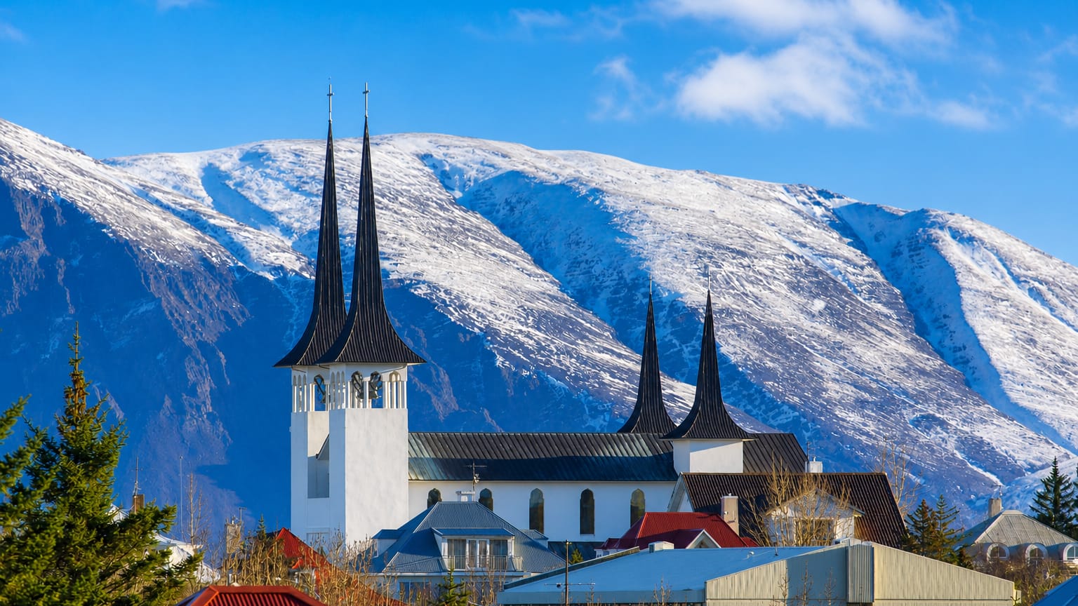 A white church with tall dark spires above Kópavogur, Iceland, set against snow-capped mountains and clear blue sky.