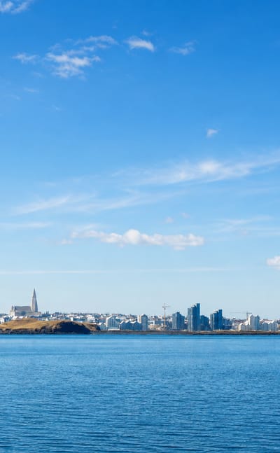 Reykjavík's low skyline across the calm blue water of Faxaflói bay, Hallgrímskirkja's spire rising under a partly cloudy sky