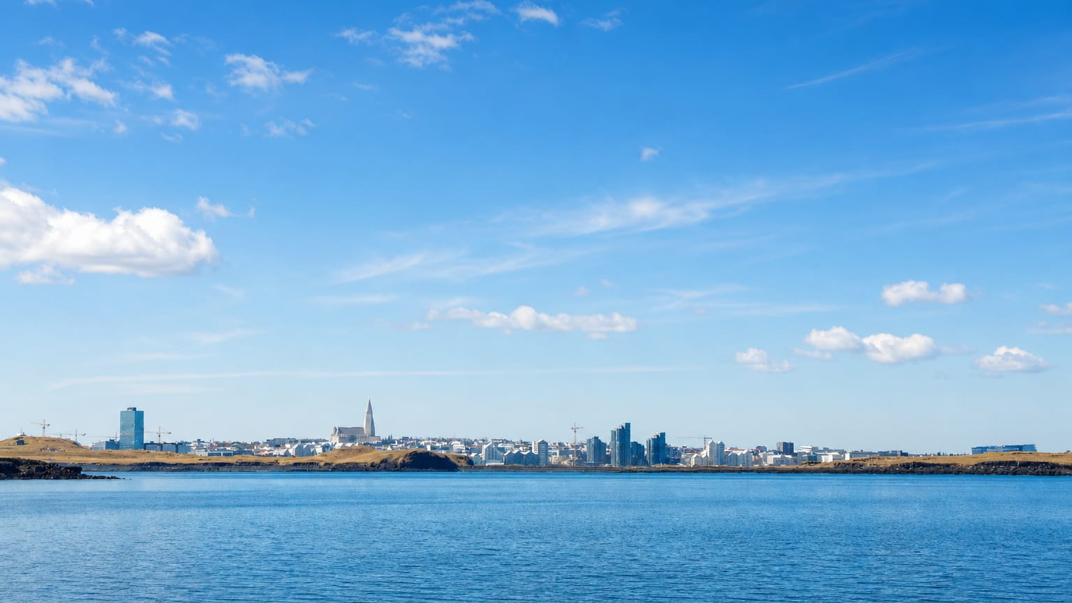 Reykjavík's low skyline across the calm blue water of Faxaflói bay, Hallgrímskirkja's spire rising under a partly cloudy sky