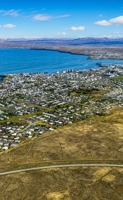 Aerial view of Keflavík, Iceland, a low-rise coastal town spread along Faxaflói bay with distant snow-capped mountains.