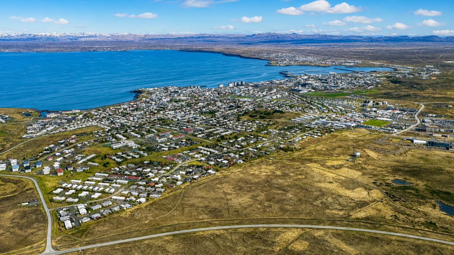 Aerial view of Keflavík, Iceland, a low-rise coastal town spread along Faxaflói bay with distant snow-capped mountains.