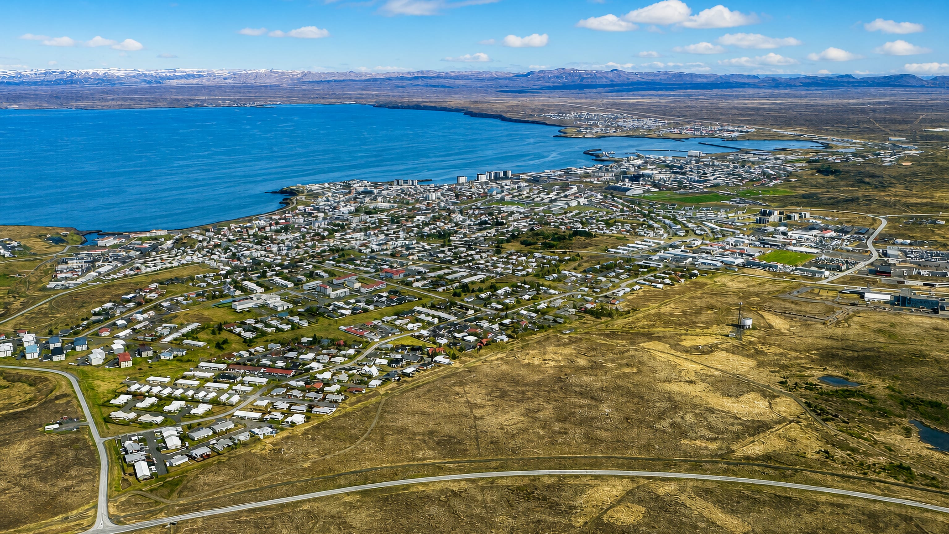 Aerial view of Keflavík, Iceland, a low-rise coastal town spread along Faxaflói bay with distant snow-capped mountains.