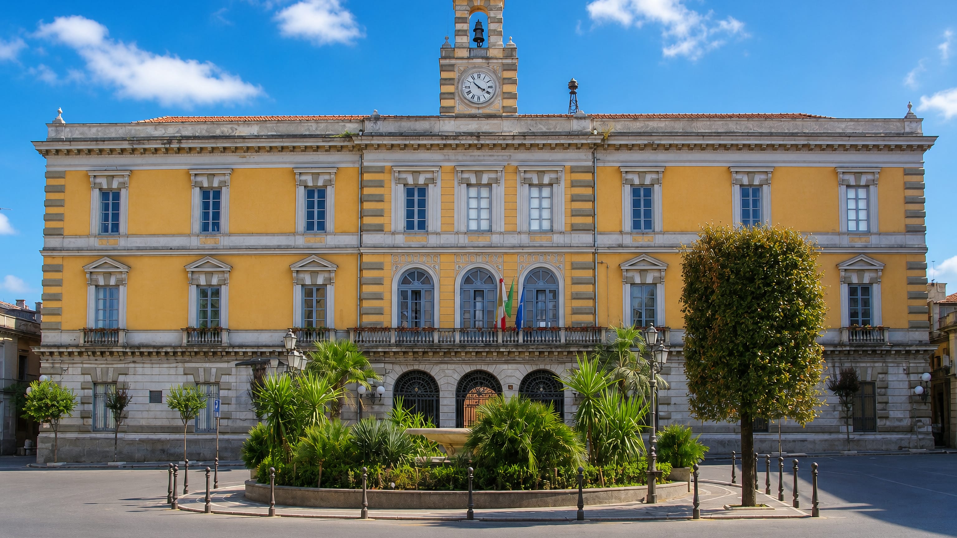 Yellow neoclassical town hall with clock and bell tower facing a palm-planted square in Afragola, Campania.