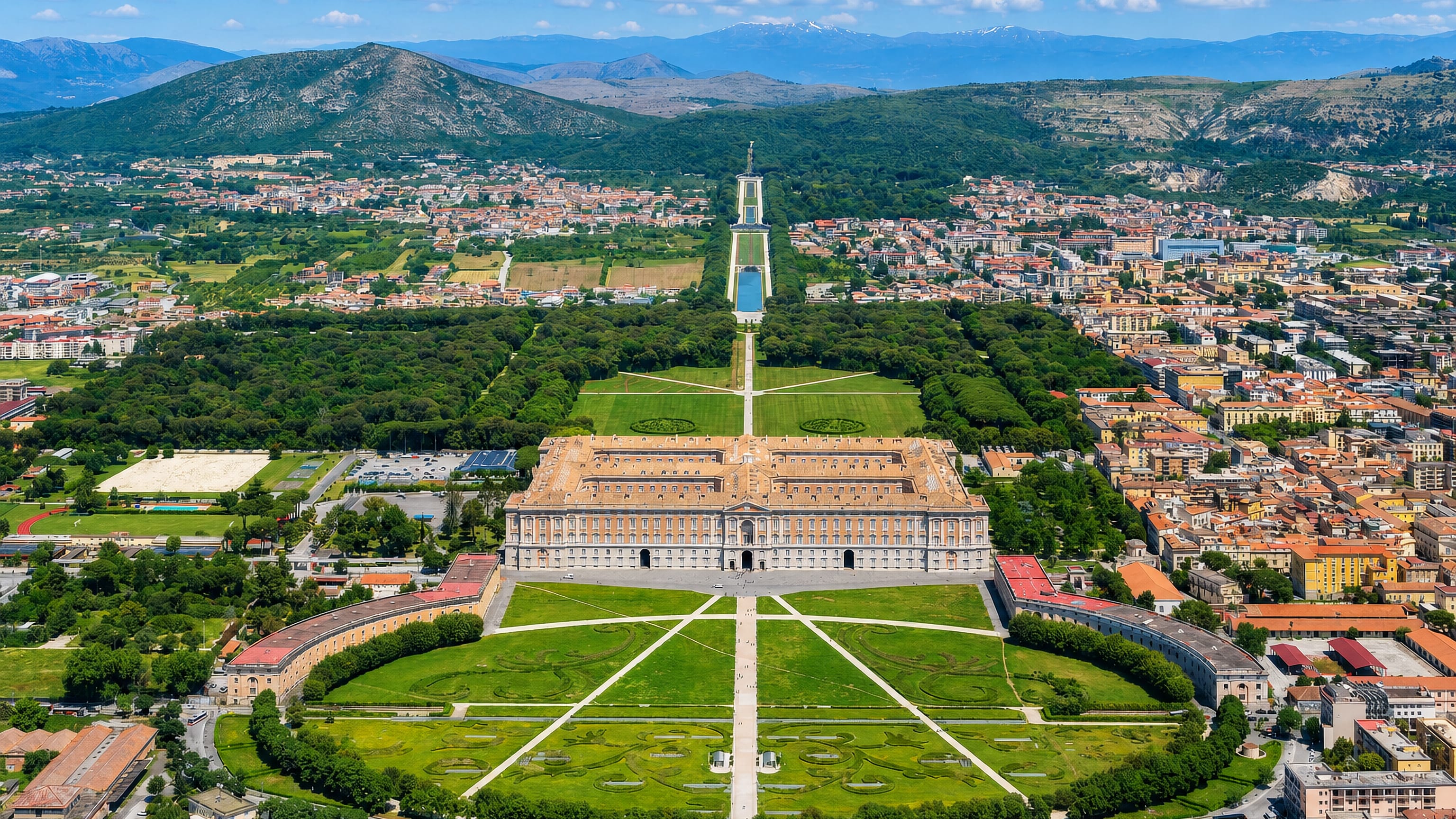 The Royal Palace of Caserta and its formal gardens from above, with the Italian town and Apennine foothills beyond.