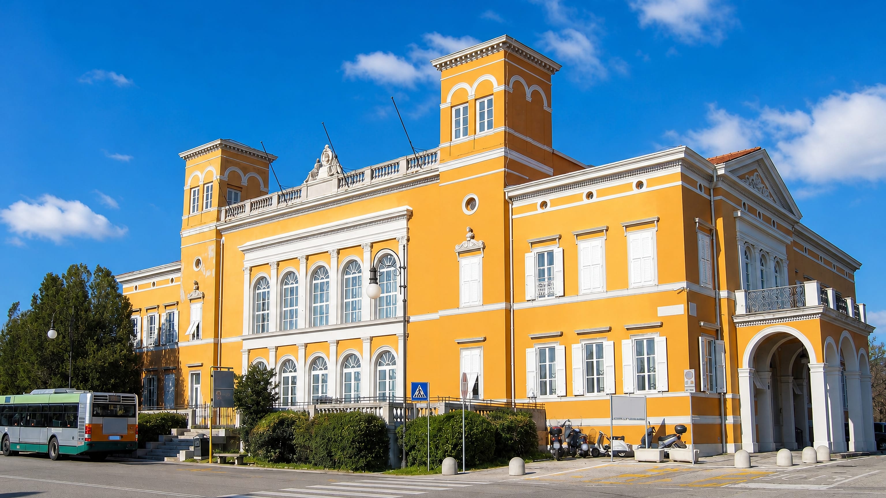 Ochre Habsburg-era villa with corner towers on a sunlit street corner in Chiadino, Trieste.