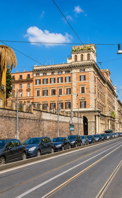 Tree-lined avenue in Rome's Esquilino district with a grand ochre palazzo, tram tracks and parked cars under blue sky.