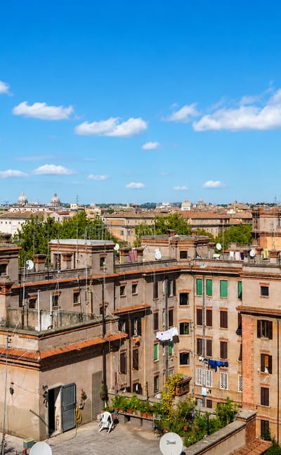 Rooftop view over Rome's Testaccio district showing terracotta-tiled buildings, satellite dishes and distant church domes.