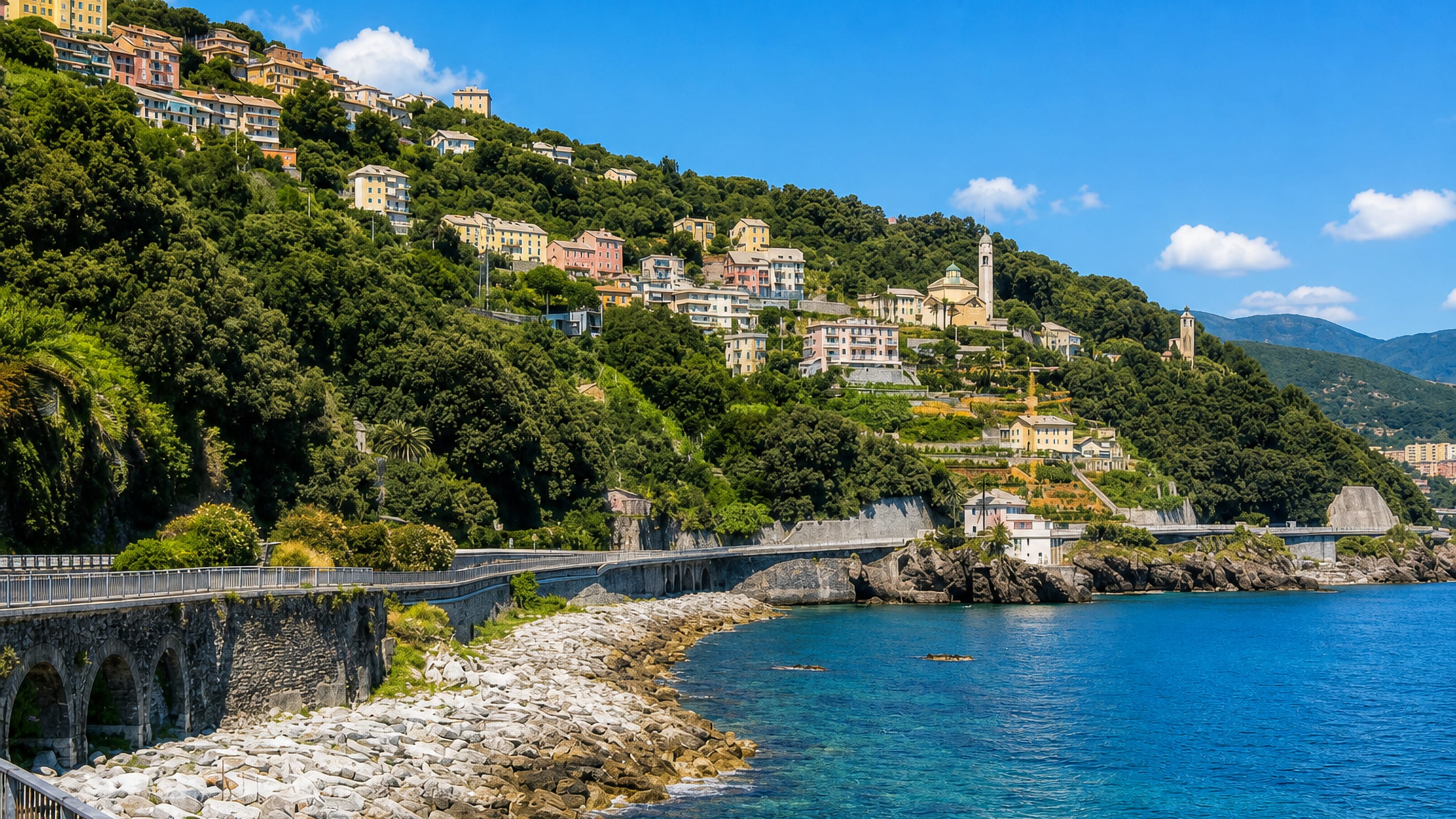 Pastel houses climbing the green hillside above the rocky coast road at Crevari, western Genoa.