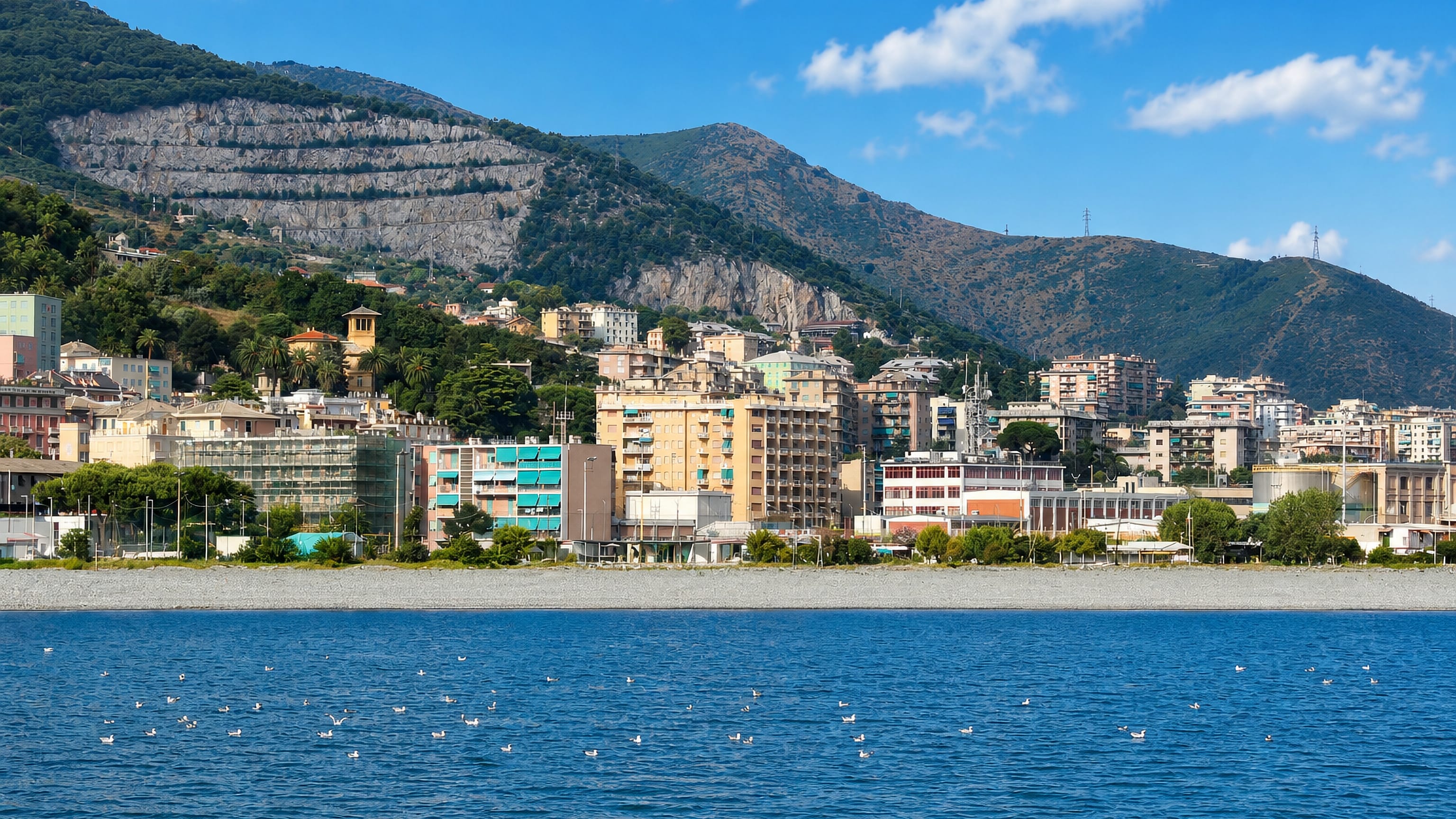 Multedo's seafront apartment blocks and pebble shore below Genoa's quarry-scarred hills, with fuel tanks at right.