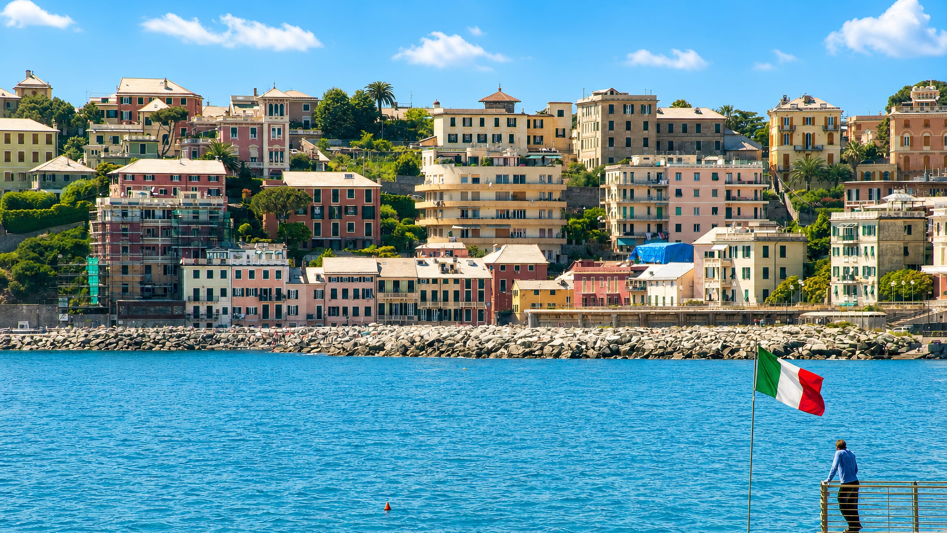 Pastel Ligurian houses above the rocky shoreline at Vernazzola in Genoa, with an Italian flag on the pier.